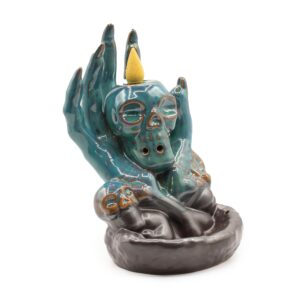 Un arzător ceramic pentru tămâie în formă de mână mistică de culoare turcoaz închis care ține un craniu mic, cu fum alb care curge în jos spre bază.